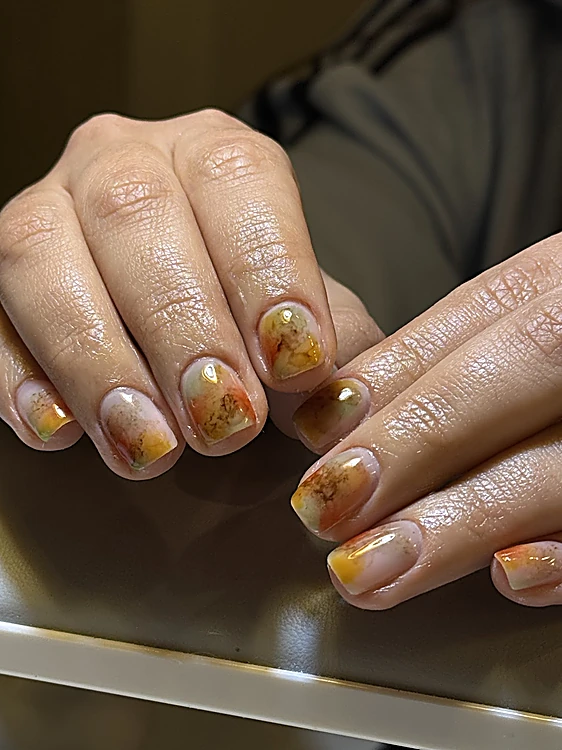 Manos con uñas nude y diseño ámbar pintado a mano, apoyadas sobre la mesa blanca del estudio con sombras largas de lámpara.