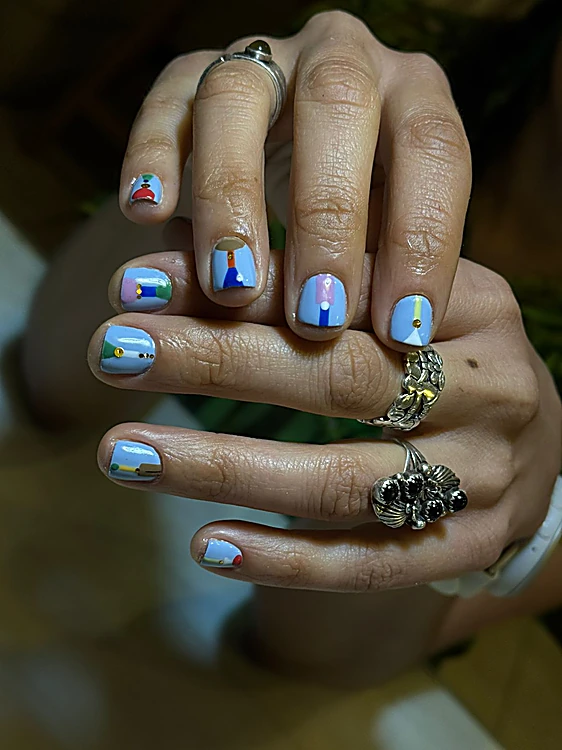 Mano con anillos de plata y uñas azul cielo con pequeñas figuras de colores pintadas a mano.