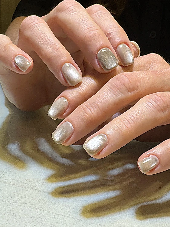 Manos con uñas cortas en tono champán perlado, con la sombra de la mano sobre la mesa.