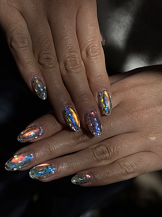 Uñas almendradas con lámina holográfica que refleja tonos azules y dorados, sobre fondo oscuro.