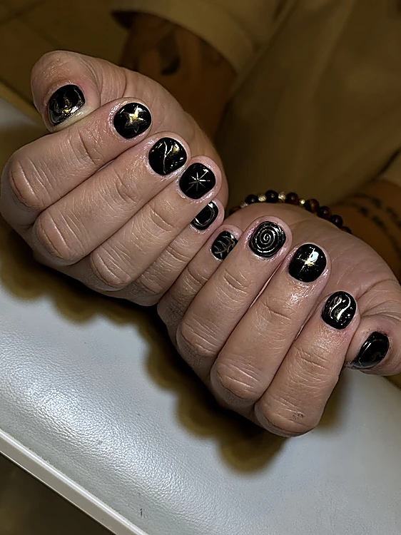 Puños con uñas cortas negras brillantes con estrellas, espirales y trazos en cromo dorado y plateado.