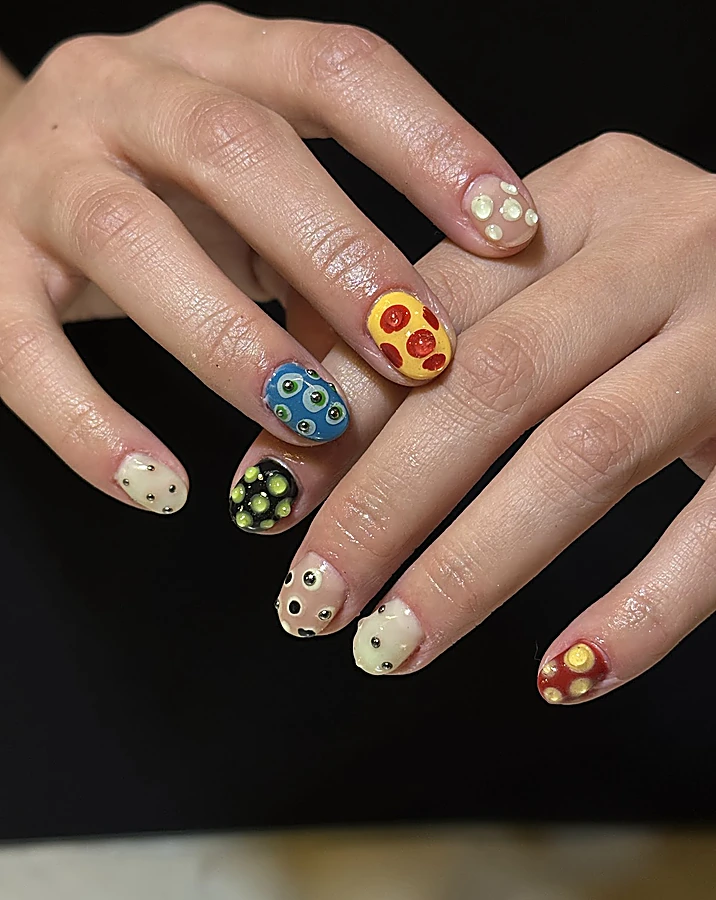 Dos manos con uñas cortas de nail art en relieve: una amarilla con rodajas de pepperoni, una azul con ojitos, una negra con puntos verdes y otras nude con perlas, sobre fondo negro.